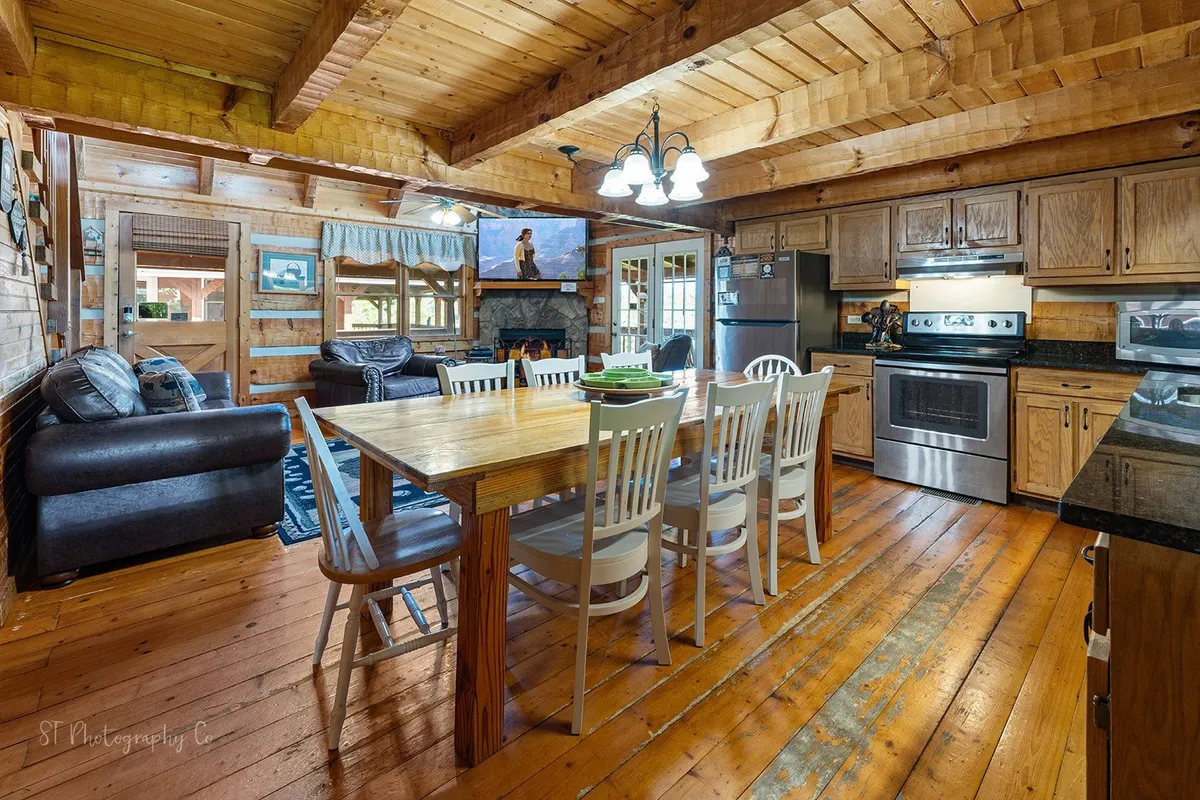 A cozy interior scene blending a large dining table, a glimpse of kitchen appliances, a welcoming seating area, and rustic wooden beams overhead. The space exudes warmth and flexibility, accommodating both dining and relaxation.