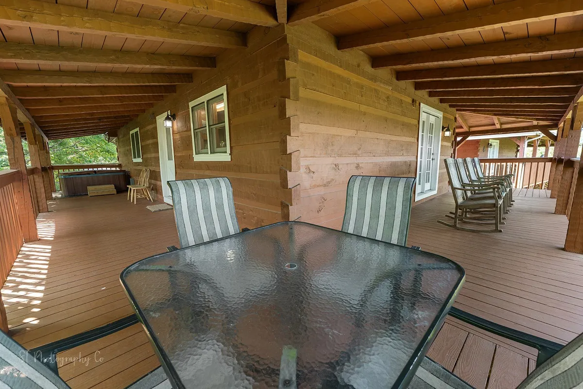 An inviting cabin deck featuring a glass table, wooden railing, rocking chairs, and large windows that reflect the surrounding trees. The space is perfect for gatherings, dining, or enjoying a sunrise with views of the natural landscape.