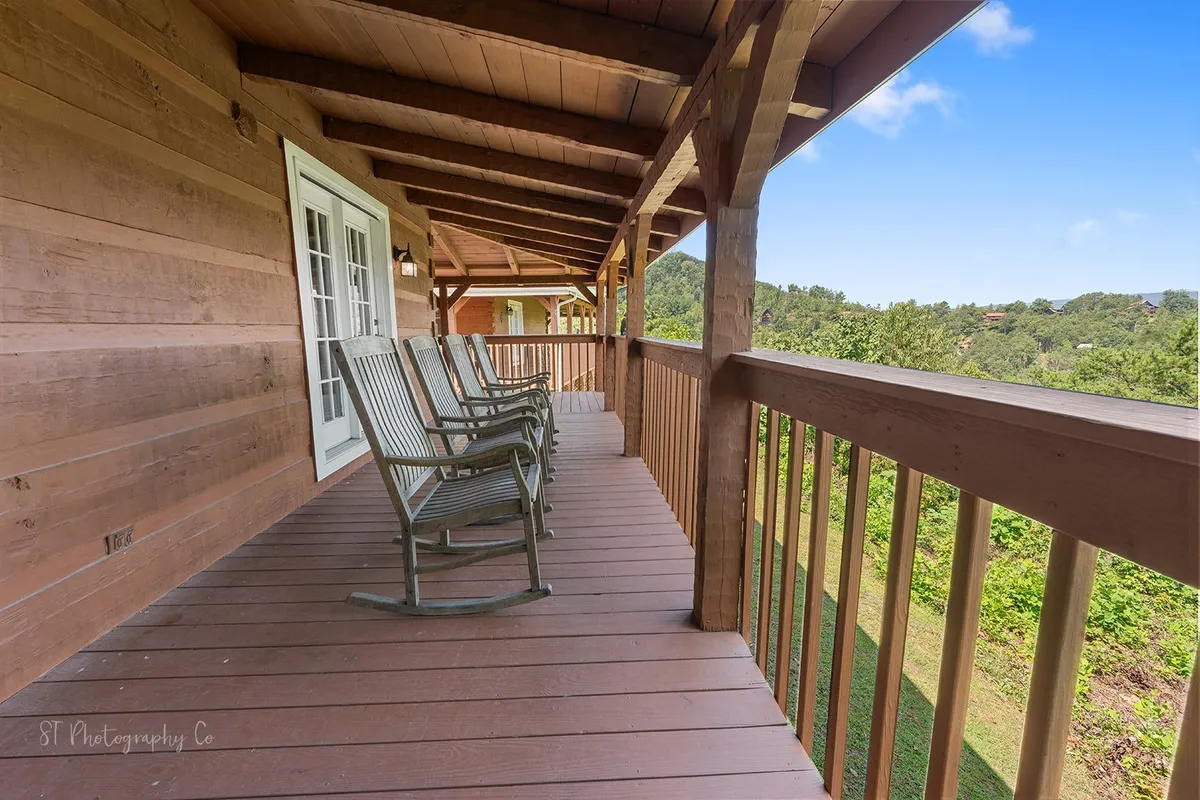 A rustic deck with classic wooden rocking chairs overlooks a sweeping, sunlit valley and distant forested hills. The spacious porch is perfect for sipping coffee, reading a book, or simply soaking in the peaceful mountain views, inviting guests of all ages to spend time together outdoors.