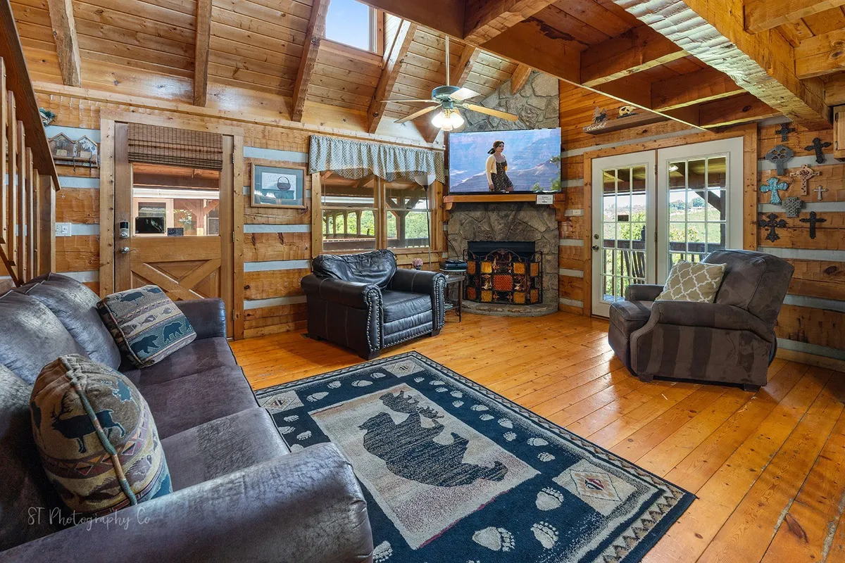 A spacious living room with a rustic stone fireplace as the centerpiece, flanked by a large TV and wooden beams overhead. Plush sofas with cozy cushions surround a bear-print rug, inviting guests to gather around. Large windows let in natural light, enhancing the warm, welcoming mood.