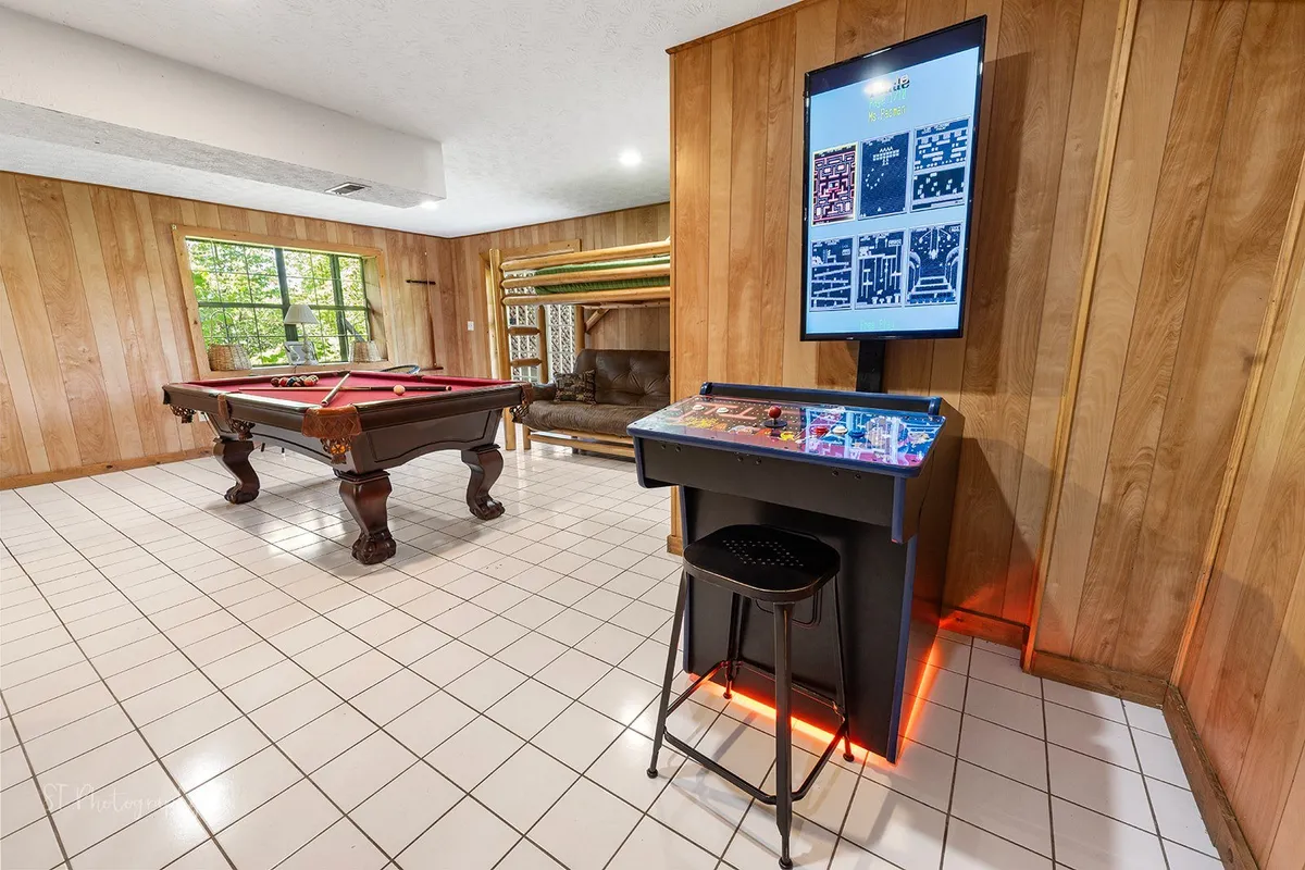This image showcases a lively game room featuring a pool table, an arcade game station, rich wood paneling, and a large window letting in plenty of natural light. There is also a comfortable couch for relaxing and socializing.