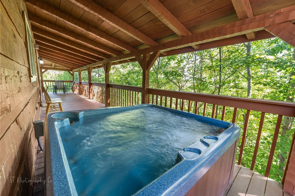This image features a steaming hot tub situated on a spacious wooden deck, surrounded by lush trees that hint at the tranquil forest setting. The deck is open to the outdoors, creating an inviting space for guests to relax and connect with nature while soaking under the stars or enjoying morning coffee.