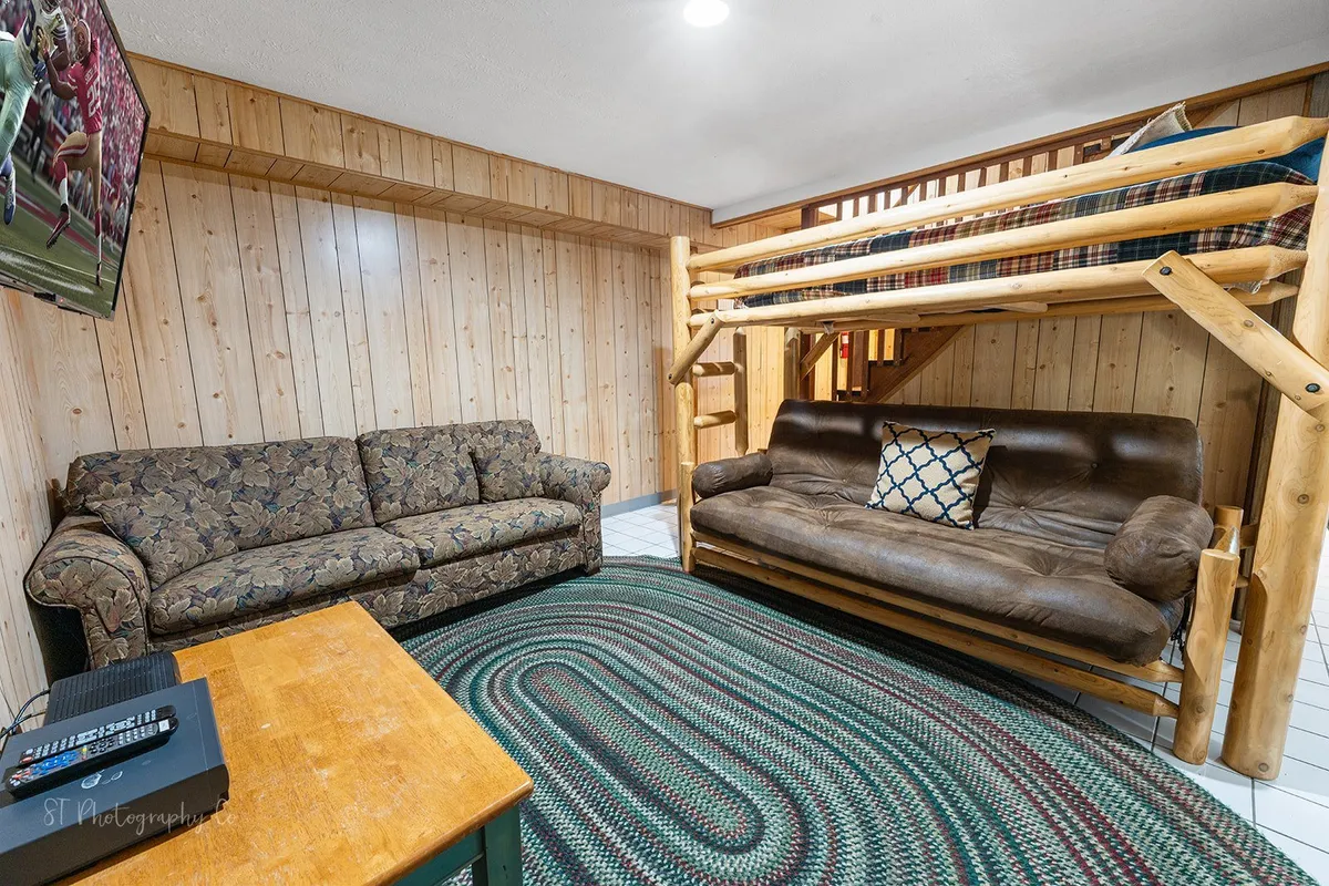 A cozy loft space featuring a wooden loft bed, floral-patterned sofa, brown leather couch, and a TV, all set against inviting wood finishes. The bright, sun-filled area creates a relaxing retreat perfect for unwinding or gaming.