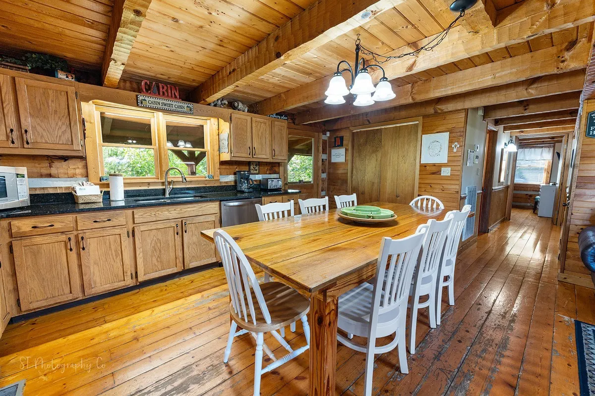 A spacious dining area featuring a large wooden table surrounded by white chairs, adjacent to kitchen cabinetry and framed by expansive windows that flood the room with natural light. The setting is ideal for family meals, group gatherings, or collaborative work sessions.