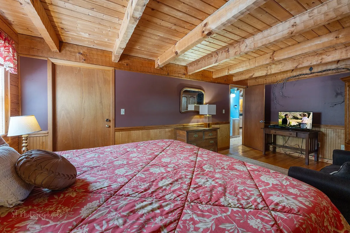 A spacious bedroom with a large bed dressed in floral bedding, wooden beams overhead, a wall-mounted television, and a decorative mirror. Soft sunlight streams in, highlighting the inviting warmth and comfort of the space.