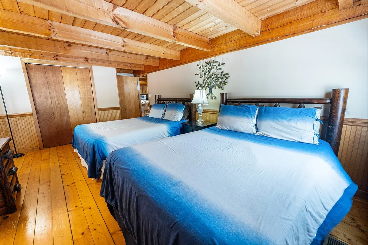 A welcoming bedroom featuring two beds with blue ombre bedding, rustic wooden beams, a bedside lamp, and a decorative plant. The space is airy and serene, suitable for families or friends sharing the room.
