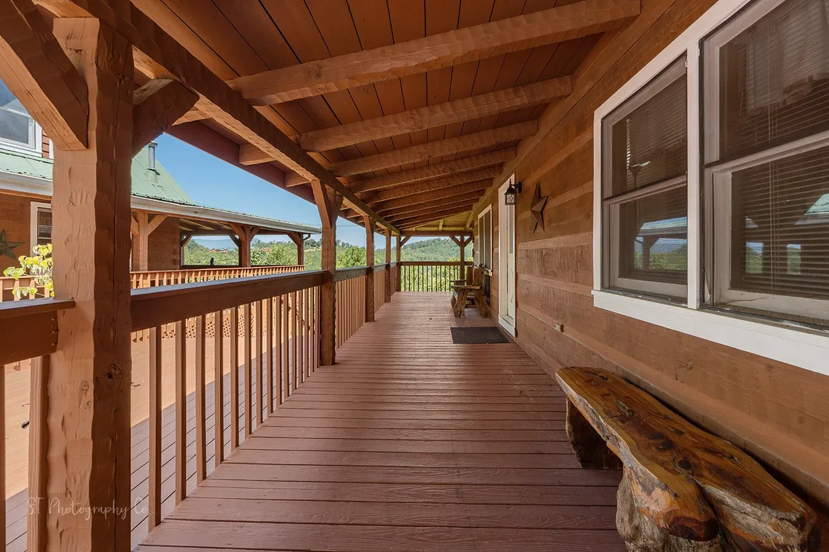 A sheltered wooden deck is framed by solid beams and railings, opening to panoramic views of rolling hills and vibrant greenery. The space exudes tranquility and invites guests to gather for conversation or quiet contemplation while immersed in the peaceful landscape.