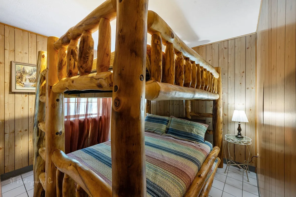 A warm and inviting bunk room with a log frame bunk bed, striped bedding, a nightstand with lamp, and rustic wood paneling. The cozy ambiance makes it ideal for children or friends looking to share a space.
