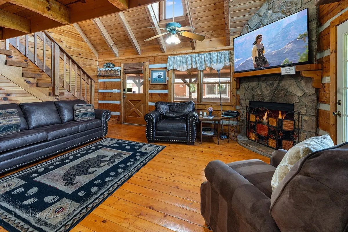 A second living area featuring comfortable leather sofas, a stone fireplace, a bear-themed area rug, and a wooden staircase leading up. The room includes a television and warm wood tones throughout, creating an inviting ambiance perfect for relaxing or socializing.